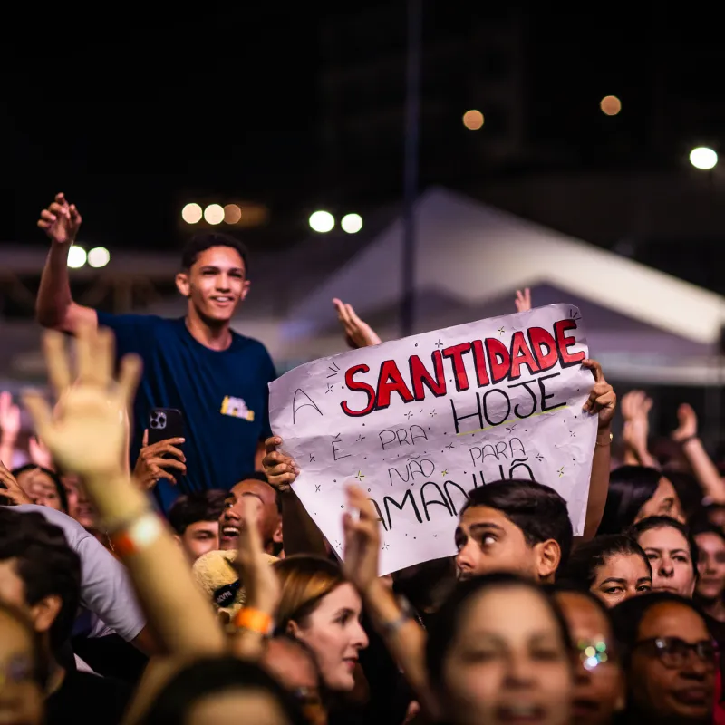 Jovem erguendo um cartaz escrito "A santidade é pra hoje, não pra amanhã" no meio da multidão do Luau.